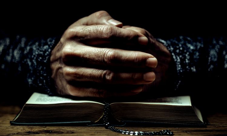 Manos entrelazadas descansando sobre un libro abierto con una hebra de cuentas, iluminación tenue y cálida en tonos oscuros