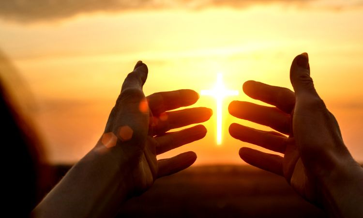 Silueta de dos manos enmarcando una cruz luminosa contra un cielo al atardecer