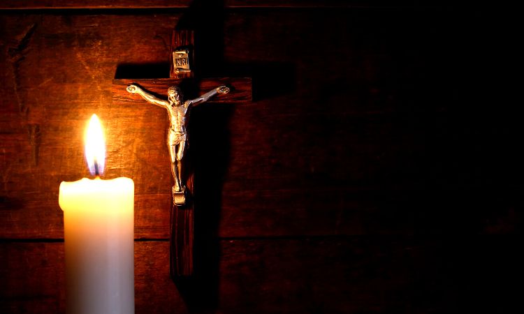 Pequeña figura metálica sobre una cruz de madera junto a una vela encendida, tonos marrones y naranjas